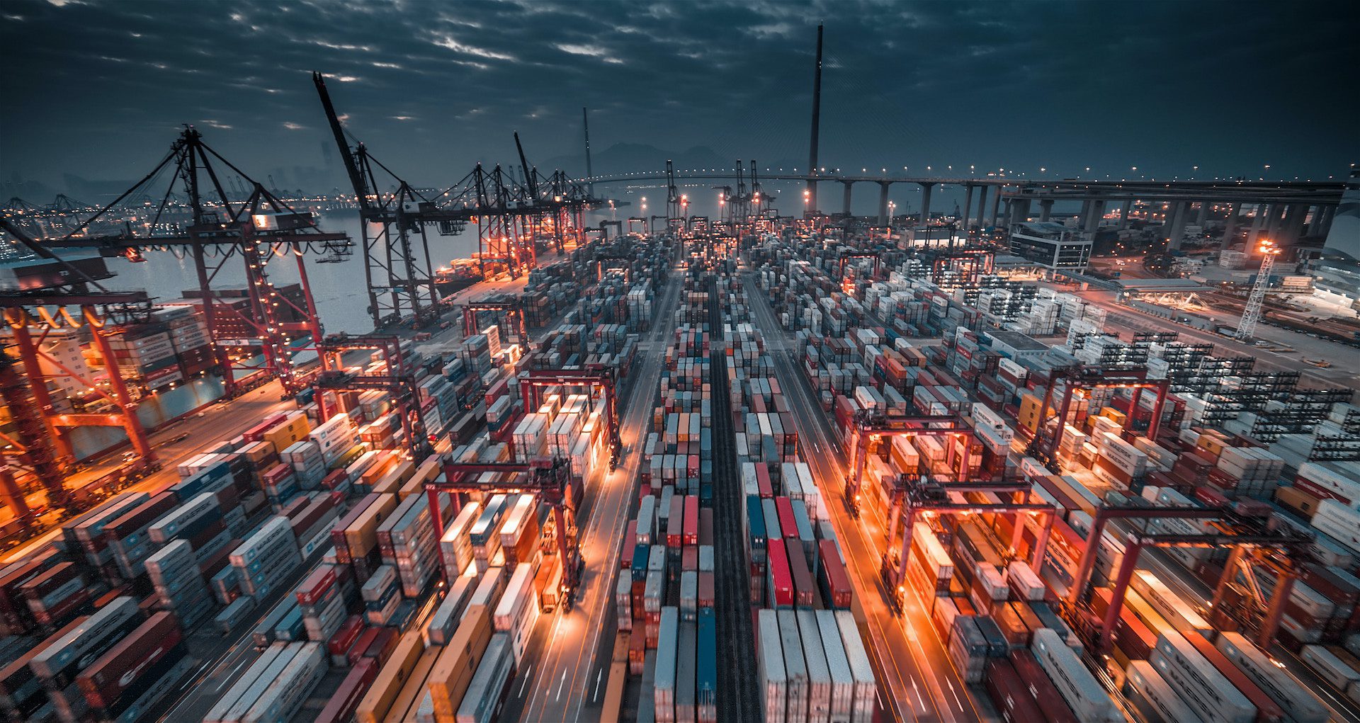 rows of shipping containers in a port at night time with a bridge in the background