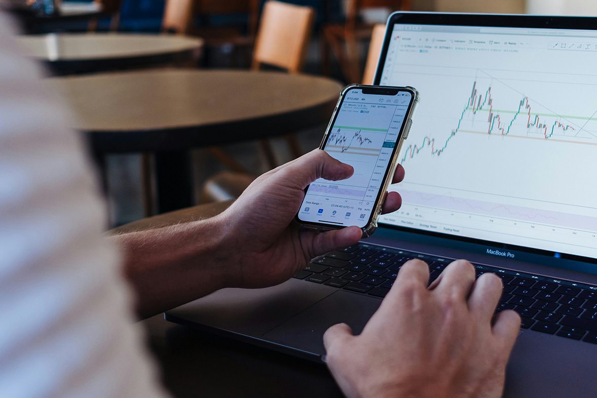 person on phone displaying a graph on screen and laptop in the background displaying a graph