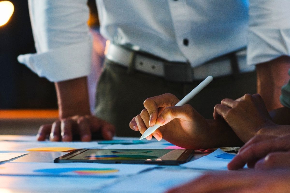 A close-up shot of hands using a stylus on a tablet device placed on a table covered with papers and charts
