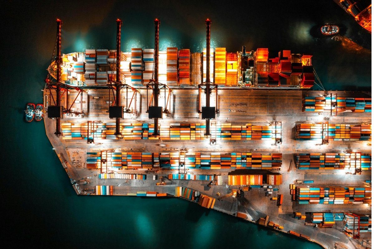 An aerial nighttime view of a busy shipping port with a large cargo ship being loaded amidst colorful shipping containers.