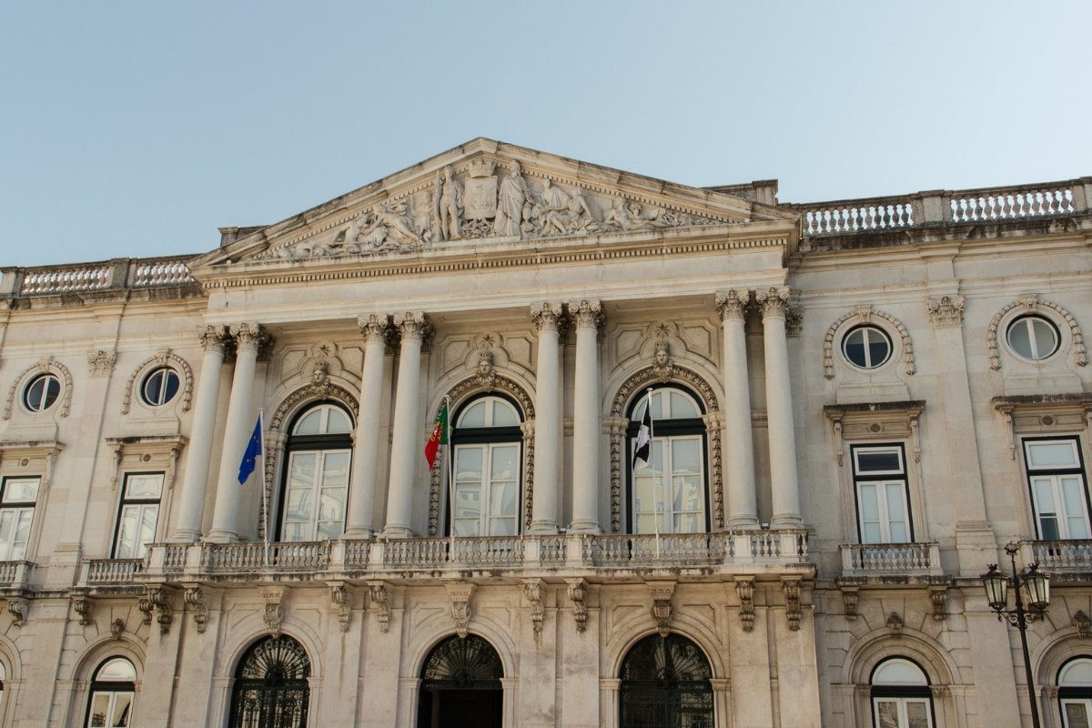 A European government building with classical architecture, featuring columns, arched windows, and decorative carvings under a clear sky