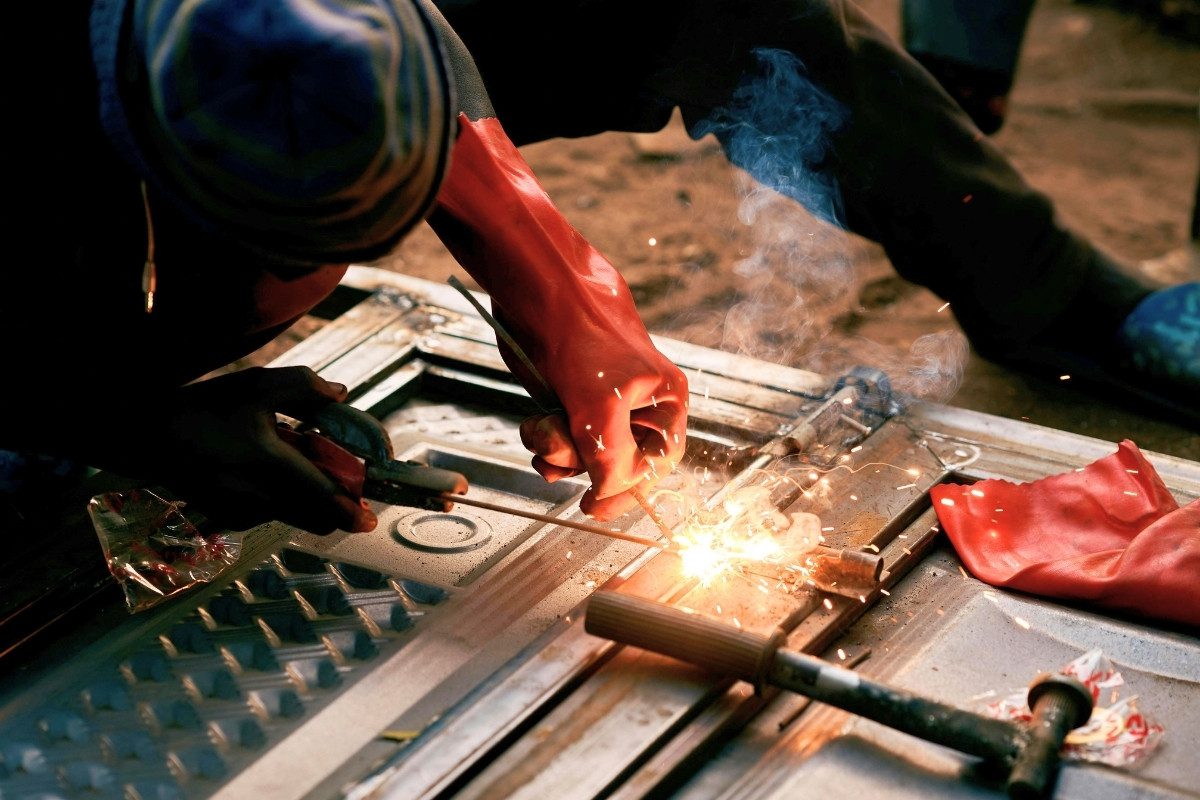 A person wearing red gloves welds metal, with bright sparks flying and smoke rising