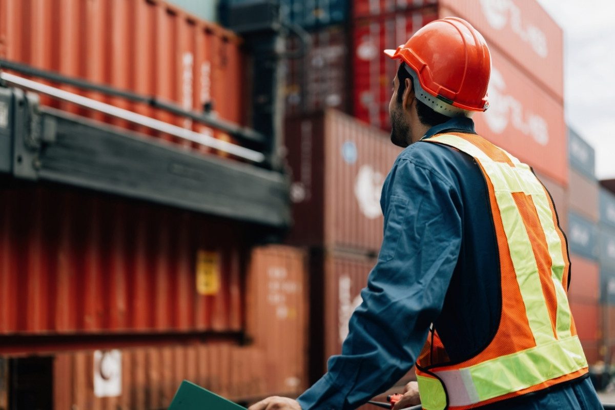 A male worker wearing a hard hat and reflective vest stands in a shipping yard looking at stacks of containers