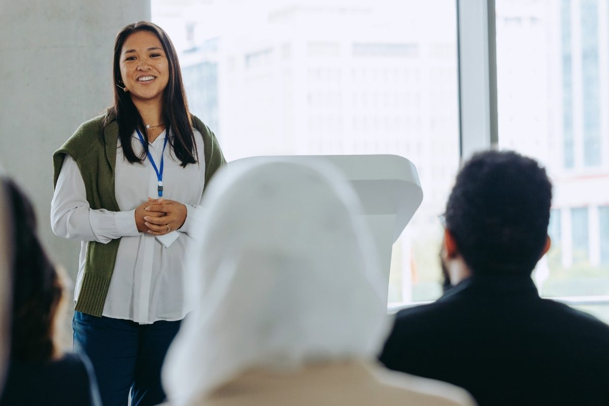 A woman speaking to an audience, smiling and gesturing with her hands