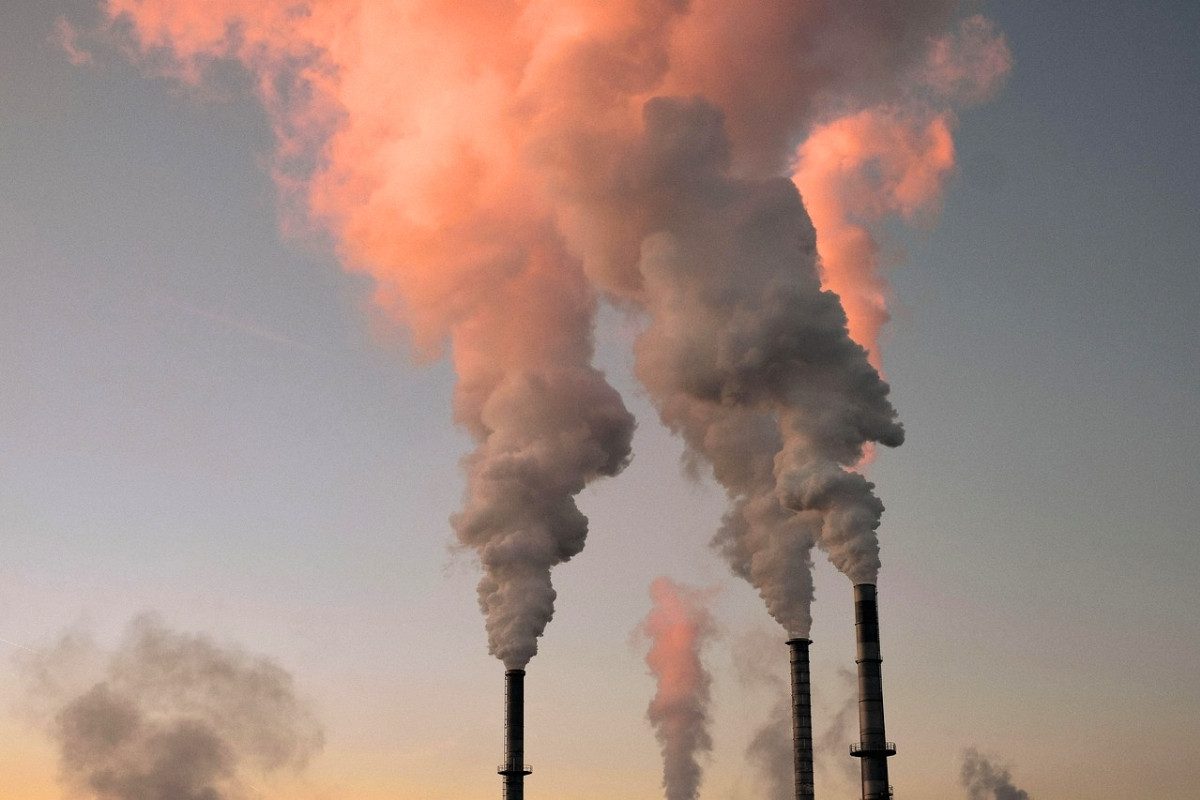Tall smokestacks emitting thick plumes of smoke against a pale sky with orange from the setting sun